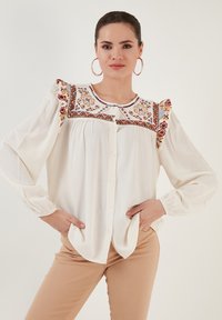 White button-up blouse with colorful embroidered accents and ruffled shoulder detail, featuring long sleeves and a relaxed fit.