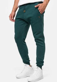 Teal joggers made of soft fabric with a slim fit, featuring side pockets and ribbed cuffs. Accented by a small logo on the thigh.