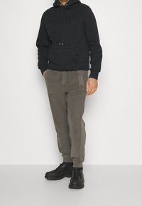 Uomo che indossa un felpa nera con cappuccio, pantaloni jogger grigi con tasche e sneaker nere, in piedi su uno sfondo bianco semplice.