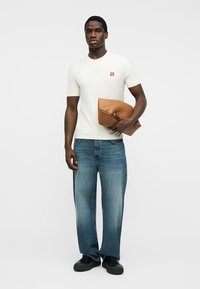 Άνδρας στέκεται φορώντας λευκό πουκάμισο με κοντό μανίκι και μικρό λογότυπο ζώου, μπλε τζιν, μαύρα παπούτσια, κρατώντας διπλωμένη καφέ δερμάτινη τσάντα κάτω από το ένα μπράτσο.