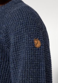 Granatowy sweter z dzianiny z teksturowanym wzorem oraz brązową skórzaną łatą w kształcie lisa na lewym ramieniu.