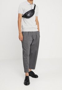 Czarna nylonowa saszetka noszona na ciele na jasnoszarym polu, w połączeniu z szarymi dopasowanymi spodniami i czarnymi sznurowanymi butami.