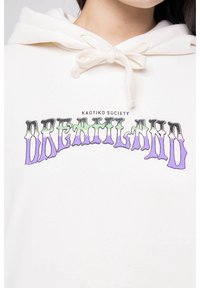 Bílá mikina s kapucí, která má stahovací šňůrku a grafický potisk. Design obsahuje slovo "DREAMLAND" v purpurové a zelené barvě s efektem kapání.
