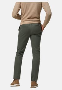 Pantalons ajustés verts en tissu lisse, avec une coupe slim et deux poches arrière. Portés avec un pull beige et des chaussures marron.