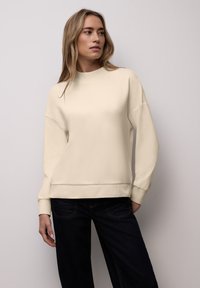 Sweat-shirt couleur crème avec un col et des poignets côtelés, présentant une coupe décontractée et une texture lisse, assorti à un jean en denim foncé.