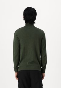 Pull à col roulé vert foncé avec des poignets et un ourlet côtelés, fabriqué dans un matériau doux. La vue montre l'arrière du vêtement. Un pantalon noir est visible.