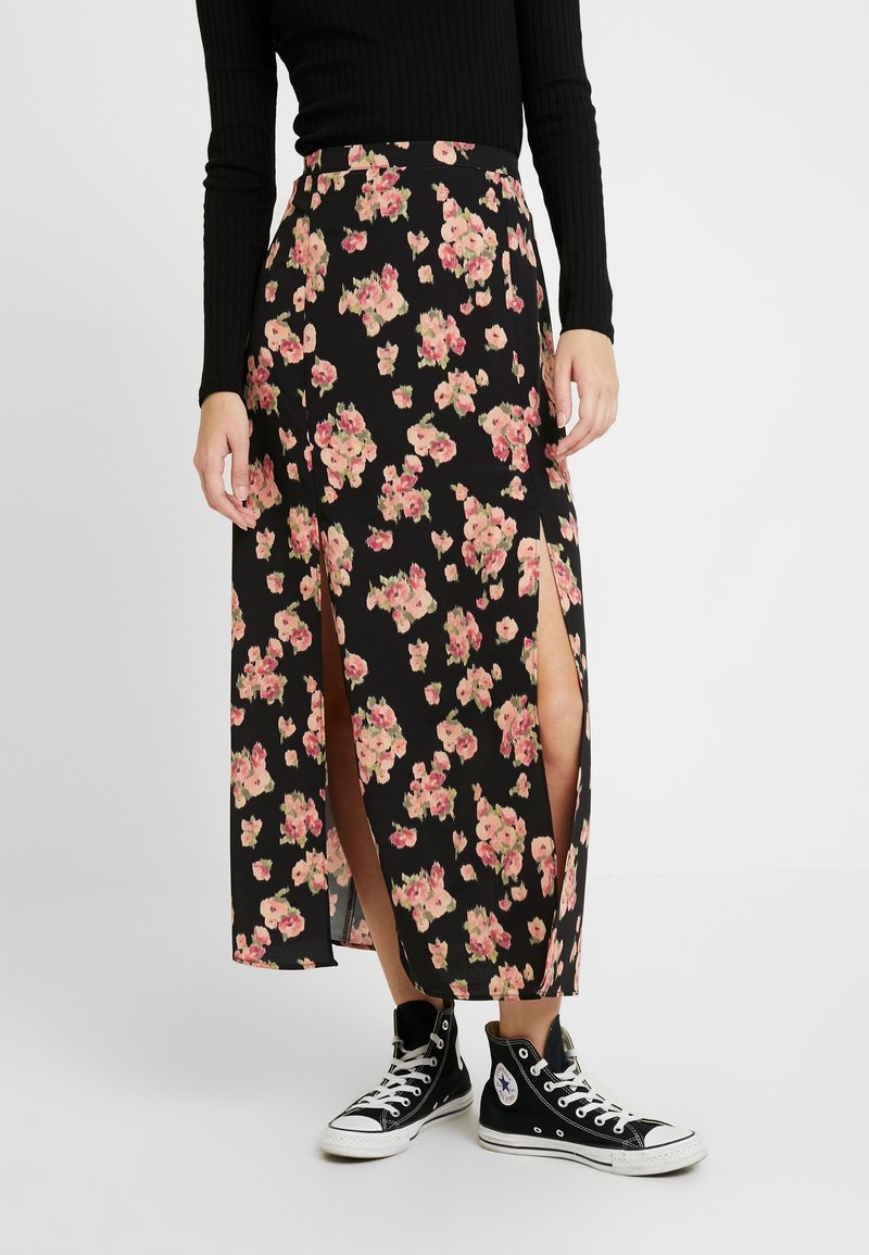 Jupe longue noire à motifs floraux avec des fentes hautes jusqu'à la cuisse, en tissu léger, ornée de fleurs roses et crème. Assortie à des baskets montantes noires.