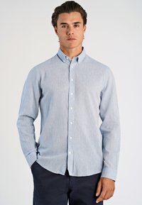 Giovane uomo che indossa una camicia azzurra chiara con colletto e pantaloni scuri, in piedi contro uno sfondo chiaro e semplice.