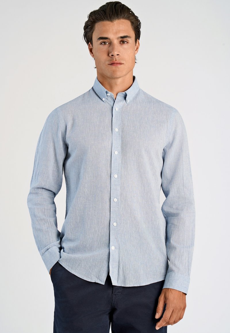 Giovane uomo che indossa una camicia azzurra chiara con colletto e pantaloni scuri, in piedi contro uno sfondo chiaro e semplice.