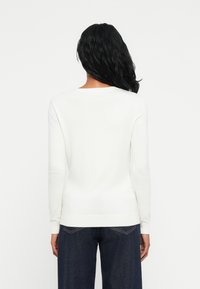 Maglione bianco a maniche lunghe con scollatura rotonda e orlo a coste. Tessuto lavorato a maglia con una texture liscia e morbida. Indossato sopra jeans scuri.