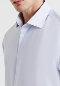 Primo piano di una persona che indossa una camicia azzurra chiara con un motivo a fine scacchi e bottoni bianchi, colletto visibile, senza accessori.