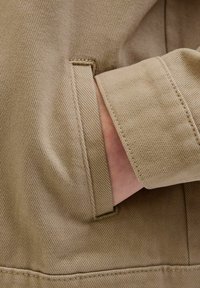 Beige pamut dzseki részlet, oldalsó zsebbel és karpánttal. Az anyag texturált felülettel rendelkezik, látható varrással.