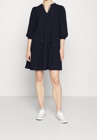 Robe bleu marine avec décolleté en V, manches bouffantes 3/4 et jupe à volants. Comprend un détail de lien avec des franges. Associée à des baskets blanches.