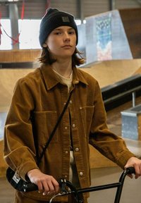 Bruine corduroy jas met twee zijzakken, gedragen over een licht shirt, in combinatie met een zwarte gebreide beanie. Een zwarte tas hangt over de schouder.