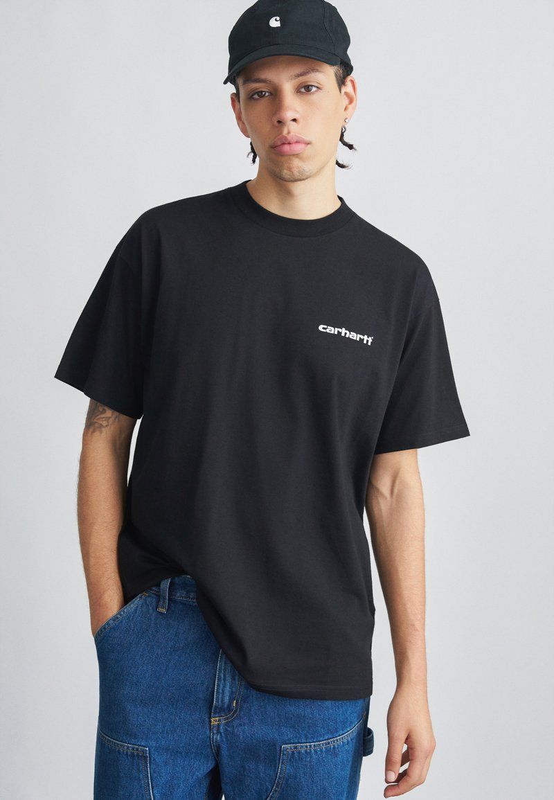 Camiseta de algodón negra con cuello redondo y mangas cortas. Presenta el logo blanco "carhartt" en el frente. Se lleva con pantalones vaqueros azules.