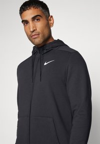 Sudadera con cremallera negra y capucha con cordón, bolsillos frontales y un logotipo de Nike en blanco en el pecho. Hecha de un material liso y ligero.