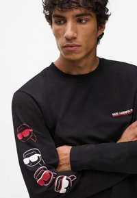 Melnā garajumta kreklā ir neliela sarkana logo uz krūtīm un krāsaini attēli ar saulesbrillēm pa kreisi no piedurknes.