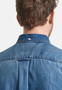 Chemise en denim de teinte bleu clair, présentant un col classique, un pli au dos, et un petit détail en bouton à la base du col.