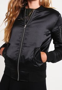 Veste bomber en satin noir avec fermeture éclair à l'avant, poignets et ourlet côtelés, poches latérales et poche zippée sur la manche. Texture lisse, design classique.