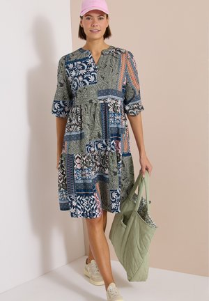 Kvinde iført en patchwork-mønstret kjole, lyserød kasket, hvide sneakers, der holder en lysegrøn quiltet taske mod en tofarvet beige væg.