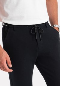 Zwarte, gestructureerde joggers met een elastische tailleband, verstelbare koordjes en zijzakken. Het model draagt een wit shirt en een zilveren horloge.