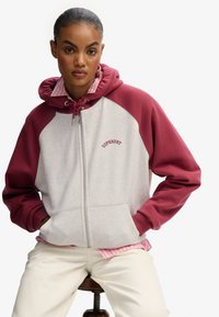 Sudadera con cremallera en gris claro con mangas raglán burdeos. Presenta una capucha con cordón y el logotipo "Superdry" bordado en el pecho. Material de mezcla de algodón.