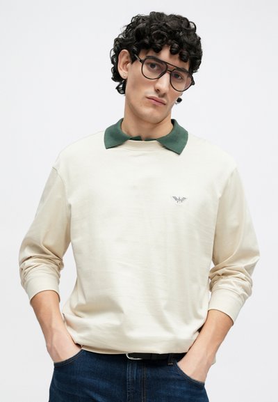 Bēša garām piedurknēm krekls ar tumši zaļu polo apkakli un logotipa uzsvaru uz krūtīm, kopā ar tumšu džinsa bikšu pāri. Gluda tekstūra.