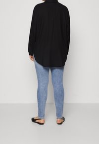 Schwarzes Langarmshirt mit abgerundetem Saum, kombiniert mit hellblauen dehnbaren Jeans und schwarzen flachen Sandalen. Glattes Material ohne Muster.