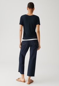 Marineblaues Kurzarmhemd mit V-Ausschnitt, kombiniert mit dunkel gewaschenen, weit geschnittenen Jeans mit fransigem Saum und zwei Gesäßtaschen. Braune Slipper.