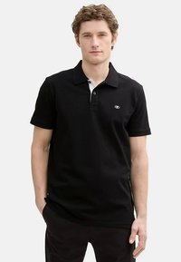 Mustast lühikeste varrukatega polo-särk, valgete kraeaktsentidega ja vasakul rinnal logoga. Valmistatud siledast, puuvillasarnasest kangast.