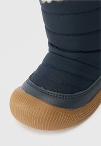 Közeli felvétel egy sötétkék téli csizmáról, amelynek párnázott szövet felső része, barna gumitalpa és a felső részen látható fehér gyapjúbélés található.