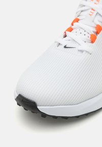 Nike sportiniai bateliai su tekstūruotu baltu paviršiumi, oranžinėmis detalėmis, perforuotomis dalimis ir juodu padu su sukibimo spygliais.