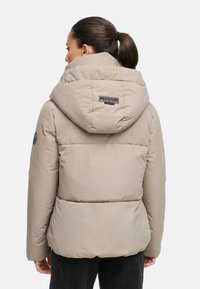 Beige pufferjacka med en stor huva, vertikal sömnadsdetalj och en etikett på övre ryggen. Materialet verkar mjukt och vadderat.