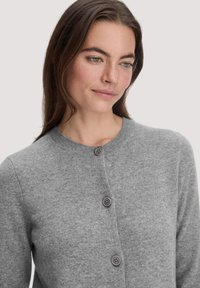 Frau mit langen braunen Haaren, die eine graue, knöpfbare Wolljacke trägt, leicht nach unten und zur Seite blickend vor einem schlichten Hintergrund.