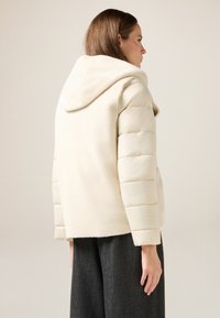 Giacca a piumino color crema con cappuccio dalla texture morbida, maniche aderenti e silhouette squadrata. Indossata sopra pantaloni scuri a gamba larga.