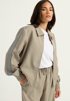 Donna che indossa una giacca beige con zip e pantaloni abbinati con dettaglio a righe bianche e nere sulle maniche, abbinata a una t-shirt bianca.