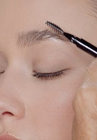 Brosse à sourcils aux poils en zigzag coiffant des sourcils naturels. Gros plan sur l'œil et la peau d'une personne, texture lisse, tons neutres, mise au point détaillée.
