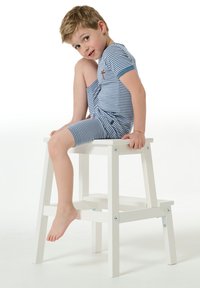 Bílý dřevěný stolička se dvěma stupni, podpírající dítě v modro-bílém pruhovaném oblečení, mírně otočené dozadu, odkryté nohy viditelné.