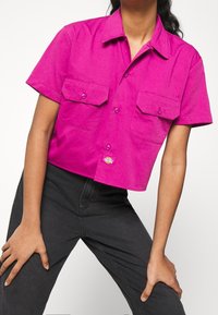 Camicia aderente a maniche corte in cotone di un brillante colore rosa, con due tasche sul petto e un design cropped, chiusura frontale con bottoni e patch logo.