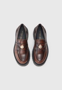 Bruine leren loafers met platte rubberen zolen, voorzien van een decoratief gouden hardwaredetail en gestikte accenten op de bovenkant.