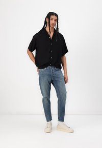 Schwarzes kurzärmeliges Hemd mit Knopfverschluss, hellblaue distressed Jeans, cremefarbene Sneakers, stehend vor einem einfarbigen Hintergrund.