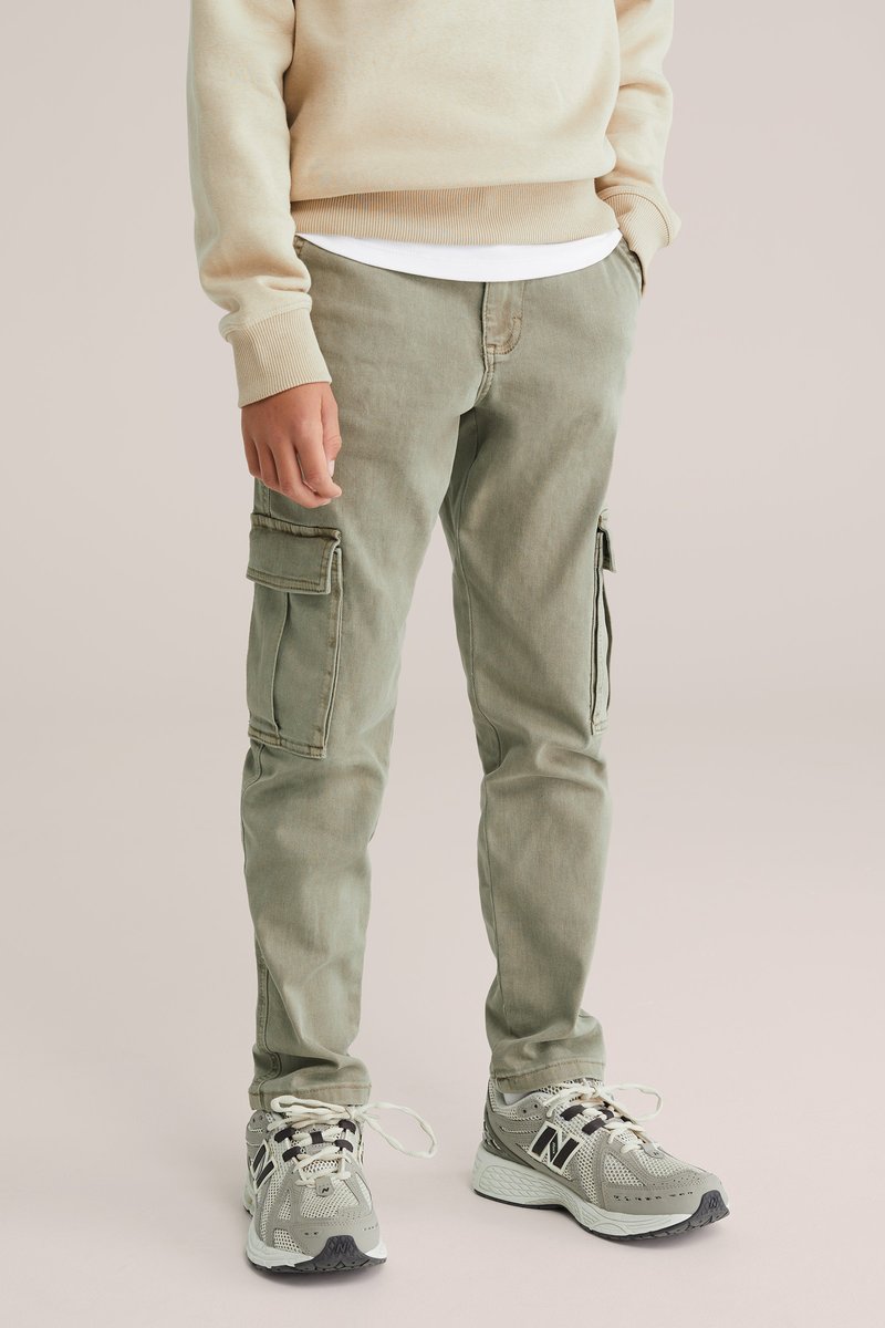 Persona llevando un sudadera beige, pantalones cargo oliva claros con bolsillos laterales y sneakers grises de New Balance sobre un fondo neutro.