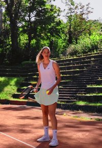 Kvinnelig tennisspiller i en hvit ermeløs topp og en lys grønn skjørt, holder en racket og to gule tennisballer på en leirgård.