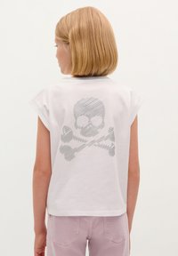 White cotton t-shirt with cap sleeves, featuring a grey skull and crossbones graphic on the back, paired with light pink pants.