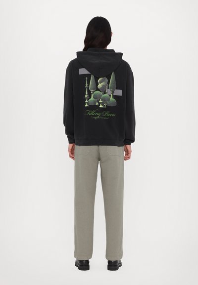 Schwarzer Hoodie mit einem grafischen Design von grünem Topiary auf dem Rücken. Kombiniert mit hellgrauen Hosen und schwarzen Schuhen, einfacher und moderner Stil.