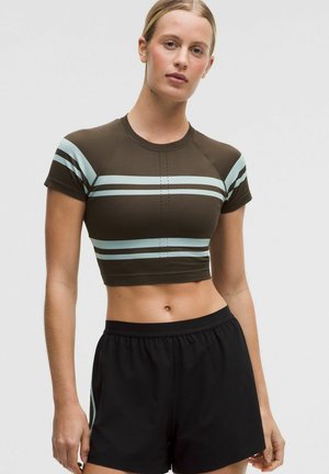 Jonge vrouw met blond haar, gekleed in een kort bruin t-shirt met lichtblauwe strepen en zwarte sportshorts, tegen een effen achtergrond.