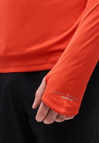 Χέρι που φοράει λαμπερό πορτοκαλί μακρυμάνικο πουκάμισο με τρύπα για τον αντίχειρα και λογότυπο «Omni-Shade», με μαύρο παντελόνι ορατό στο παρασκήνιο.