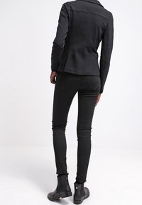 Černá přiléhavá bunda s širokými klopami, kombinovaná s černými úzkými džíny a kotníkovými botami, s hladkou texturou a minimalistickým designem.