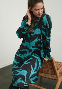 Žena sedí na tkanom drevenom kresle, má na sebe dlhé šaty s vzorom v odtieňoch teal a čiernej, pozerá dolu a má ruku blízko ucha.