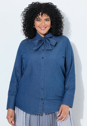 Kvinde med krøllet sort hår iført en denim-knap-op-skjorte med sløjfe ved kraven og et lysegråt plisseret skørt, smilende.